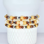 A stack of beaded bracelets in shades of gold, brown, and pearl white. The bracelets feature a variety of beads, including pearls, faceted crystals, and round beads. The bracelets are displayed on a white, textured surface.