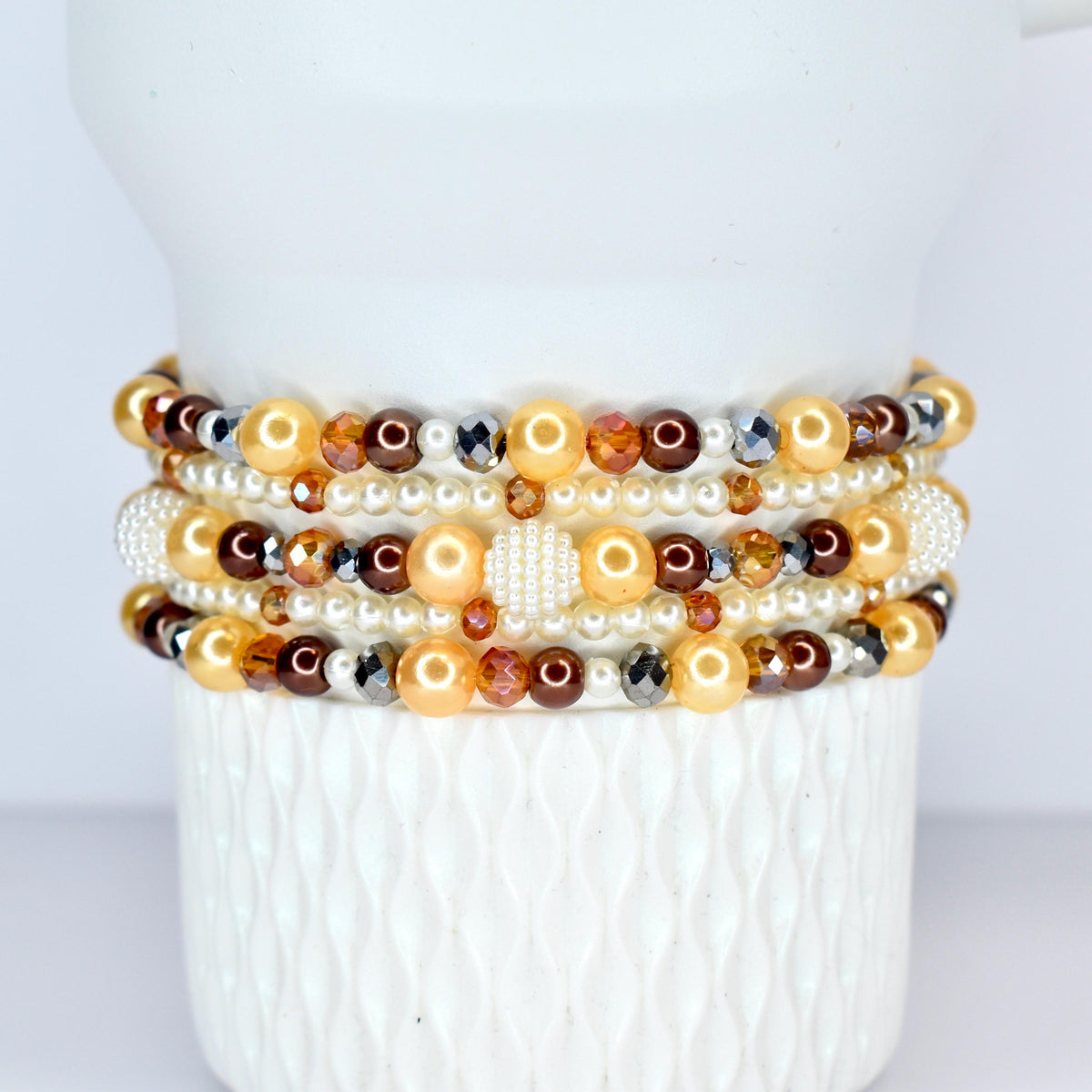 A stack of beaded bracelets in shades of gold, brown, and pearl white. The bracelets feature a variety of beads, including pearls, faceted crystals, and round beads. The bracelets are displayed on a white, textured surface.