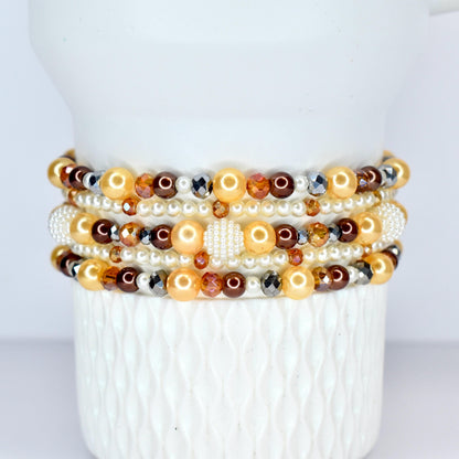 A stack of beaded bracelets in shades of gold, brown, and pearl white. The bracelets feature a variety of beads, including pearls, faceted crystals, and round beads. The bracelets are displayed on a white, textured surface.
