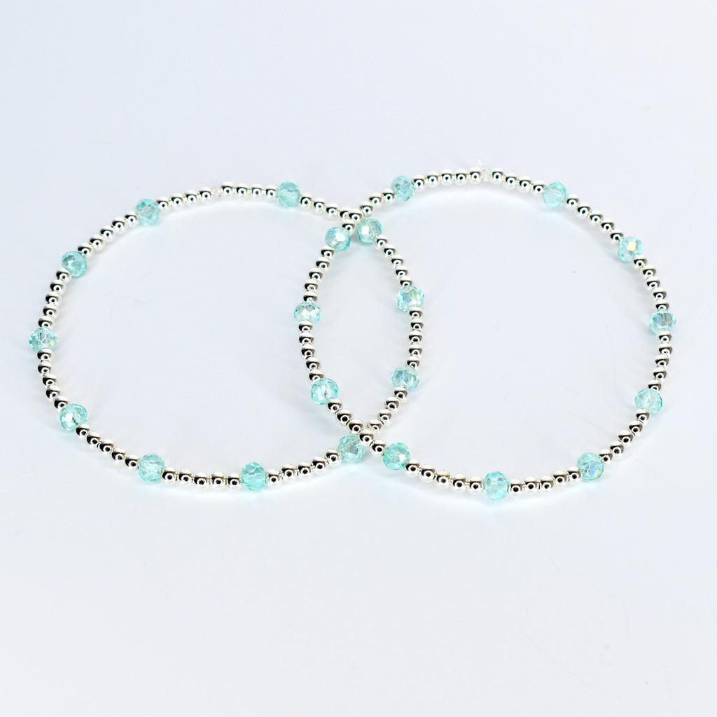 Two delicate bracelets. Each bracelet features small silver beads interspersed with larger, faceted, light blue beads. The bracelets are displayed on a white background, showcasing their simple elegance.