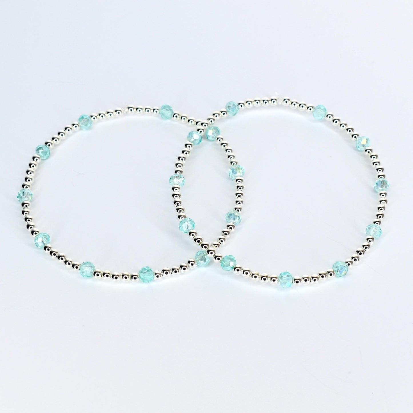 Two delicate bracelets. Each bracelet features small silver beads interspersed with larger, faceted, light blue beads. The bracelets are displayed on a white background, showcasing their simple elegance.