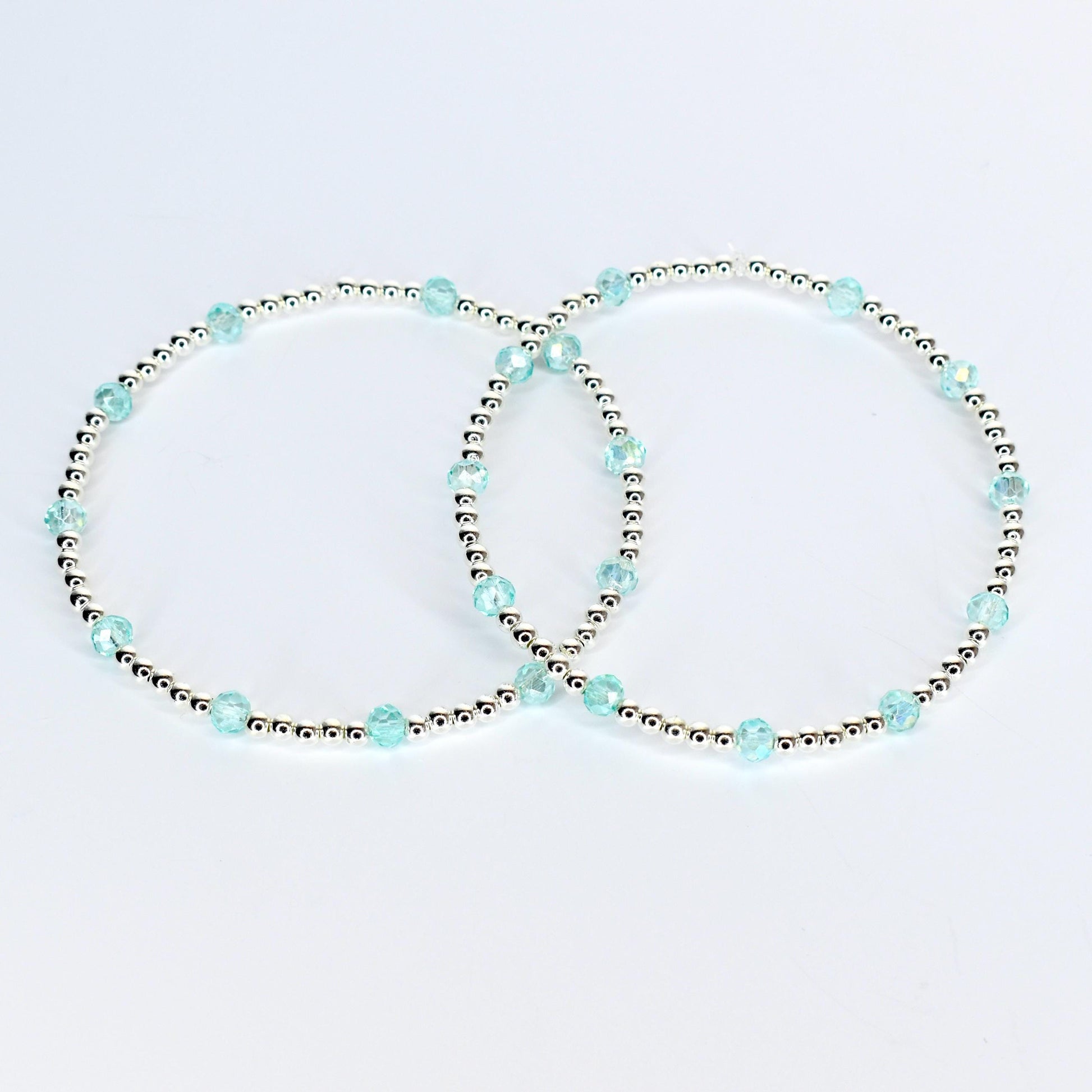 Two delicate bracelets. Each bracelet features small silver beads interspersed with larger, faceted, light blue beads. The bracelets are displayed on a white background, showcasing their simple elegance.