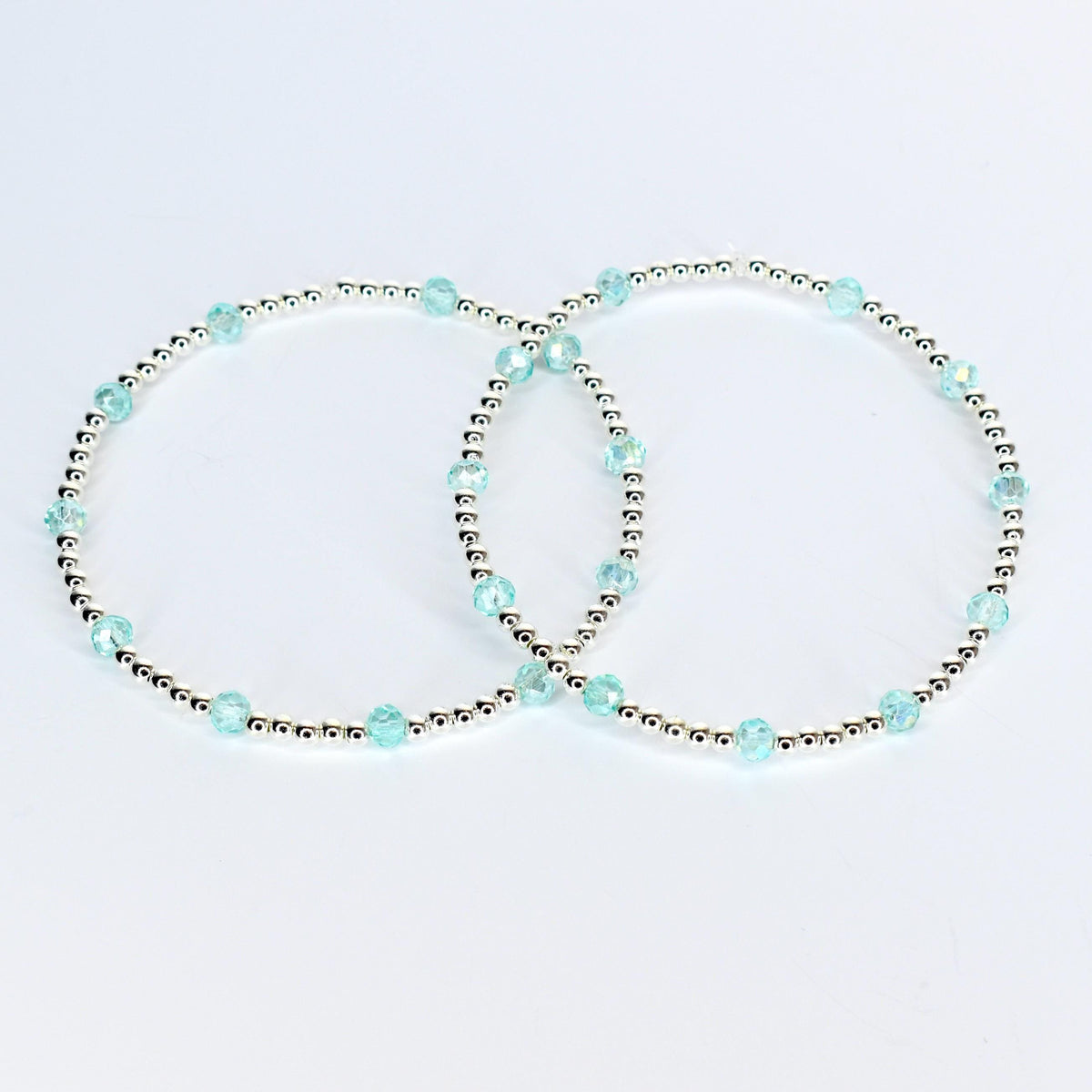 Two delicate bracelets. Each bracelet features small silver beads interspersed with larger, faceted, light blue beads. The bracelets are displayed on a white background, showcasing their simple elegance.