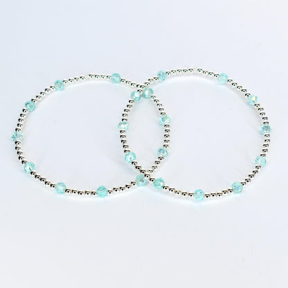 Two delicate bracelets. Each bracelet features small silver beads interspersed with larger, faceted, light blue beads. The bracelets are displayed on a white background, showcasing their simple elegance.