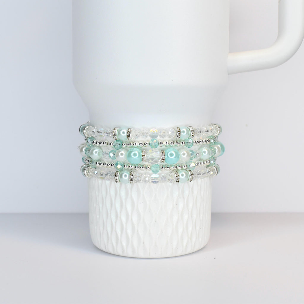 A white tumbler with a textured base is adorned with four beaded bracelets. The bracelets feature clear and light blue beads, interspersed with silver-colored spacers. The bracelets are wrapped around the tumbler.
