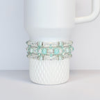 A white tumbler with a textured base is adorned with four beaded bracelets. The bracelets feature clear and light blue beads, interspersed with silver-colored spacers. The bracelets are wrapped around the tumbler.