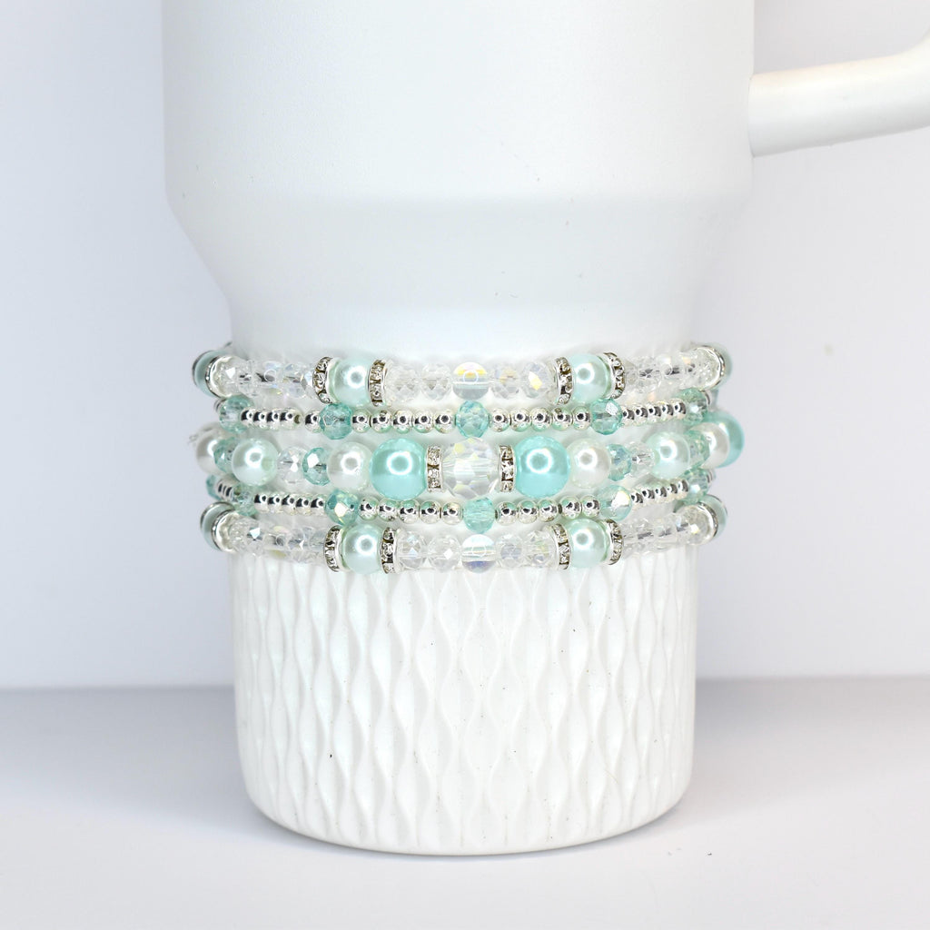 A stack of four beaded bracelets in shades of white, clear, and light blue. The bracelets feature a mix of clear beads, faux pearls, and silver-toned accents. The bracelets are wrapped around a white ceramic mug.