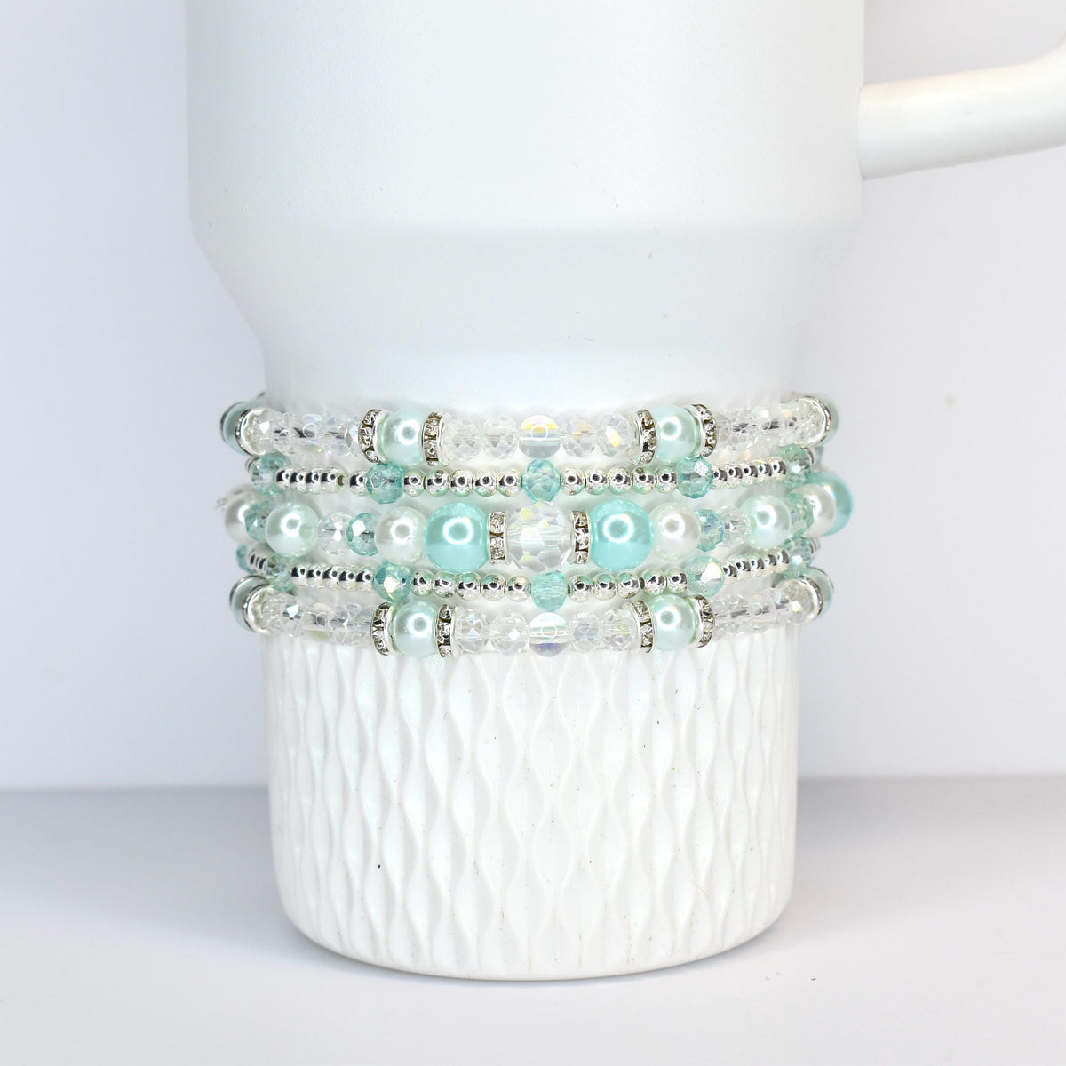 A stack of four beaded bracelets in shades of white, clear, and light blue. The bracelets feature a mix of clear beads, faux pearls, and silver-toned accents. The bracelets are wrapped around a white ceramic mug.