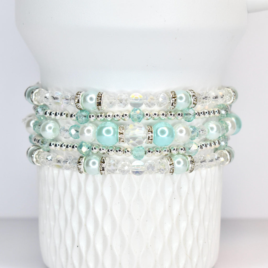 A stack of four beaded bracelets. Each bracelet features a combination of clear and light blue beads, with silver accents. The bracelets are displayed on a white, textured surface.