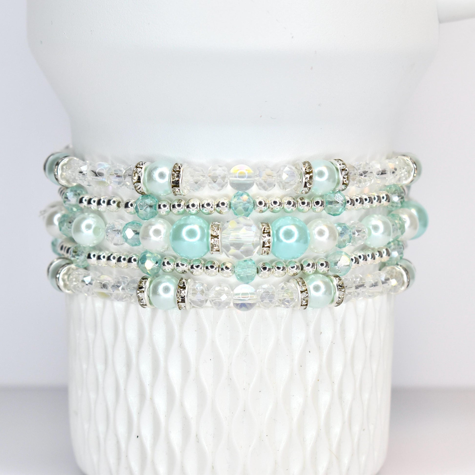 A stack of four beaded bracelets. Each bracelet features a combination of clear and light blue beads, with silver accents. The bracelets are displayed on a white, textured surface.