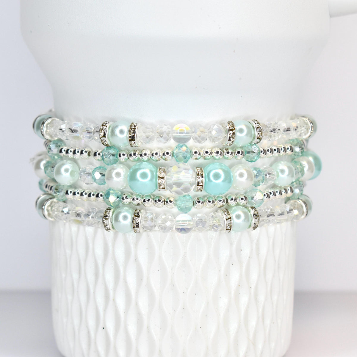 A stack of four beaded bracelets. Each bracelet features a combination of clear and light blue beads, with silver accents. The bracelets are displayed on a white, textured surface.