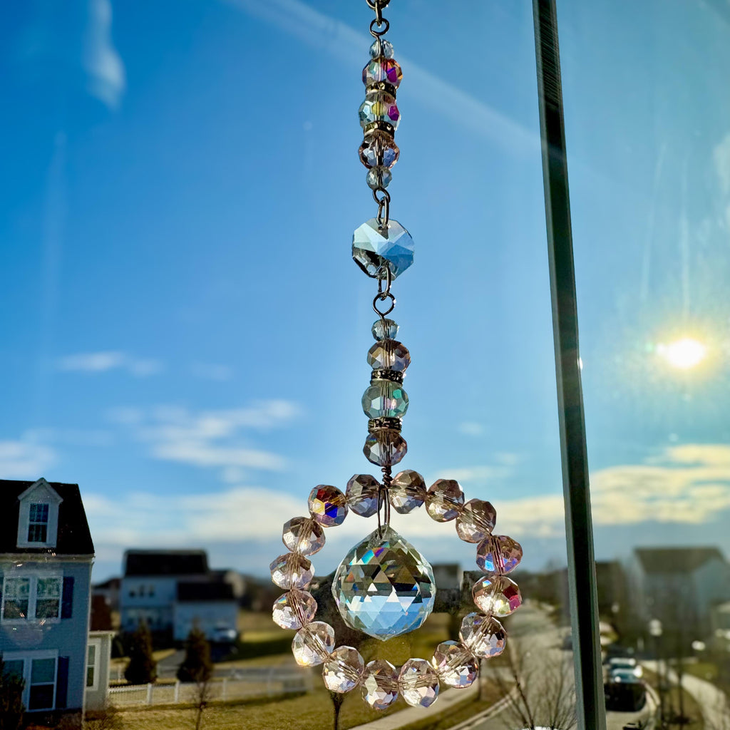 A clear crystal suncatcher with a circular design and a large, faceted crystal at the bottom. The suncatcher is suspended by a chain and smaller crystals. The background shows a blue sky and houses.