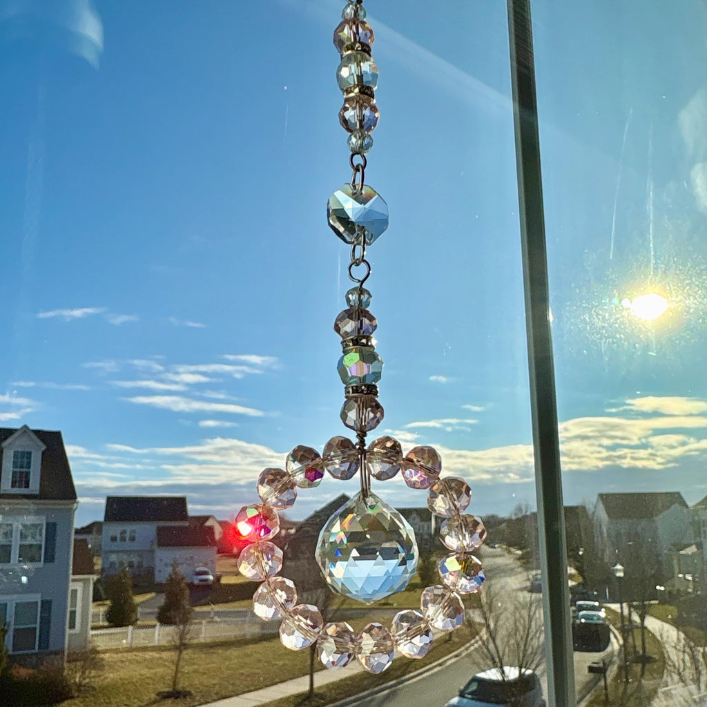 A crystal suncatcher with a circular design, hanging in front of a window. The suncatcher features a large, faceted crystal ball at the bottom, surrounded by a ring of smaller, pink-toned crystals. Above, a series of clear and iridescent crystals are strung together.