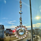 A crystal suncatcher with a circular design, hanging in front of a window. The suncatcher features a large, faceted crystal ball at the bottom, surrounded by a ring of smaller, pink-toned crystals. Above, a series of clear and iridescent crystals are strung together.
