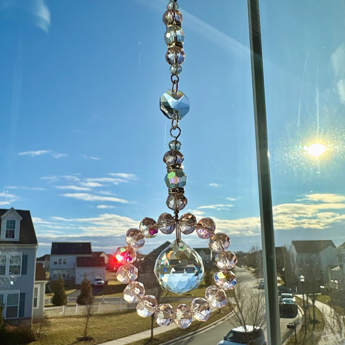 A crystal suncatcher with a circular design, hanging in front of a window. The suncatcher features a large, faceted crystal ball at the bottom, surrounded by a ring of smaller, pink-toned crystals. Above, a series of clear and iridescent crystals are strung together.