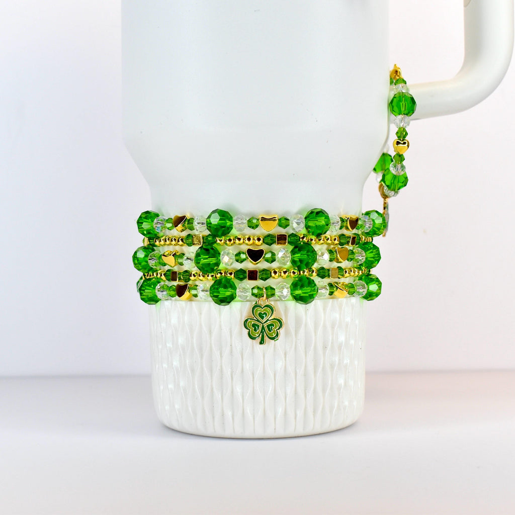 A white mug adorned with beaded bracelets in shades of green and gold. The bracelets feature round beads, gold accents, and a shamrock charm. The mug's handle is also decorated with green beads.