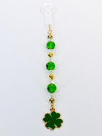 A St. Patrick's Day themed charm featuring a green four-leaf clover pendant. The charm is adorned with green and clear faceted beads, gold-colored beads, and a gold-colored chain. The charm is attached to a clear plastic loop.
