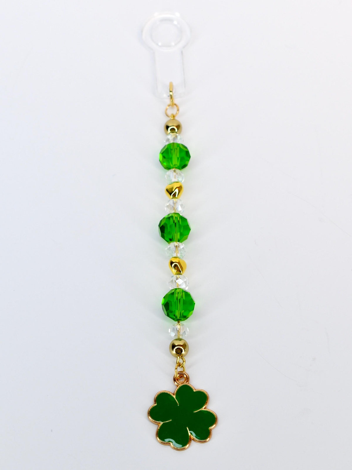 A St. Patrick's Day themed charm featuring a green four-leaf clover pendant. The charm is adorned with green and clear faceted beads, gold-colored beads, and a gold-colored chain. The charm is attached to a clear plastic loop.