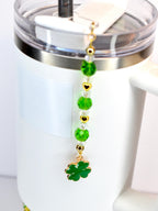 A white tumbler with a gold and green charm. The charm features green beads, gold heart accents, and a green four-leaf clover pendant. The tumbler has a clear lid and a silver band.