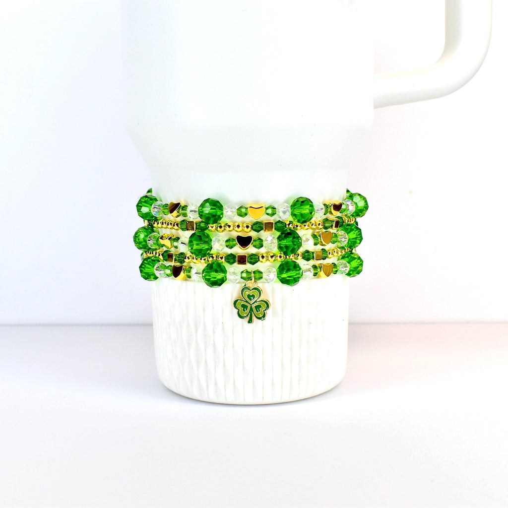 A white Stanley tumbler adorned with a St Patricks day themed beaded bracelet stack. The bracelet features green and gold beads, with a shamrock charm. The beads are arranged in multiple strands, creating a layered effect. The design is festive and suitable for St. Patrick's Day.