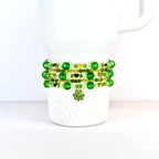 A white Stanley tumbler adorned with a St Patricks day themed beaded bracelet stack. The bracelet features green and gold beads, with a shamrock charm. The beads are arranged in multiple strands, creating a layered effect. The design is festive and suitable for St. Patrick's Day.