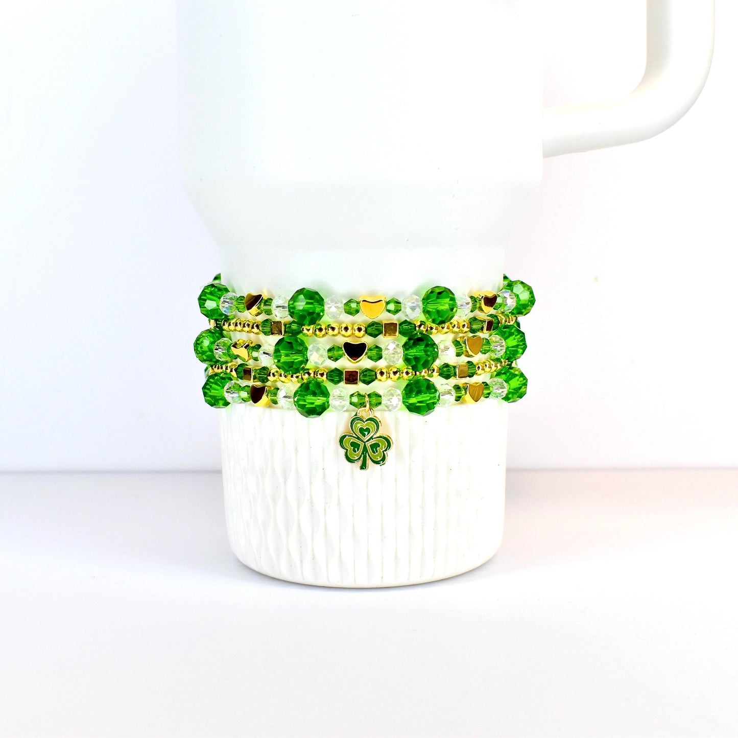 A white Stanley tumbler adorned with a St Patricks day themed beaded bracelet stack. The bracelet features green and gold beads, with a shamrock charm. The beads are arranged in multiple strands, creating a layered effect. The design is festive and suitable for St. Patrick's Day.