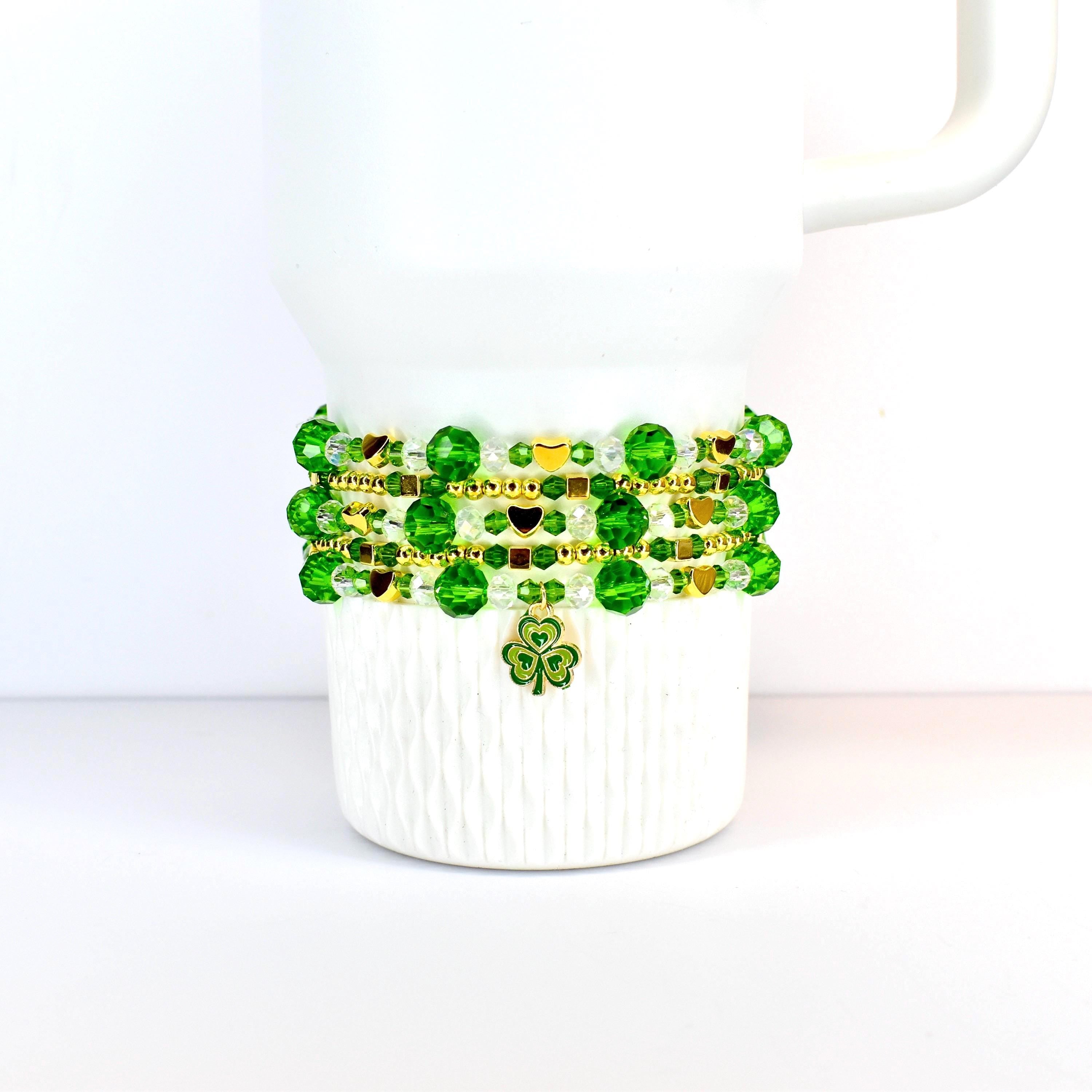 A white Stanley tumbler adorned with a St Patricks day themed beaded bracelet stack. The bracelet features green and gold beads, with a shamrock charm. The beads are arranged in multiple strands, creating a layered effect. The design is festive and suitable for St. Patrick's Day.
