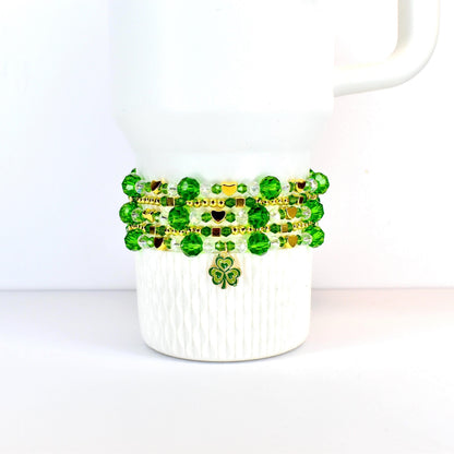 A white Stanley tumbler adorned with a St Patricks day themed beaded bracelet stack. The bracelet features green and gold beads, with a shamrock charm. The beads are arranged in multiple strands, creating a layered effect. The design is festive and suitable for St. Patrick's Day.