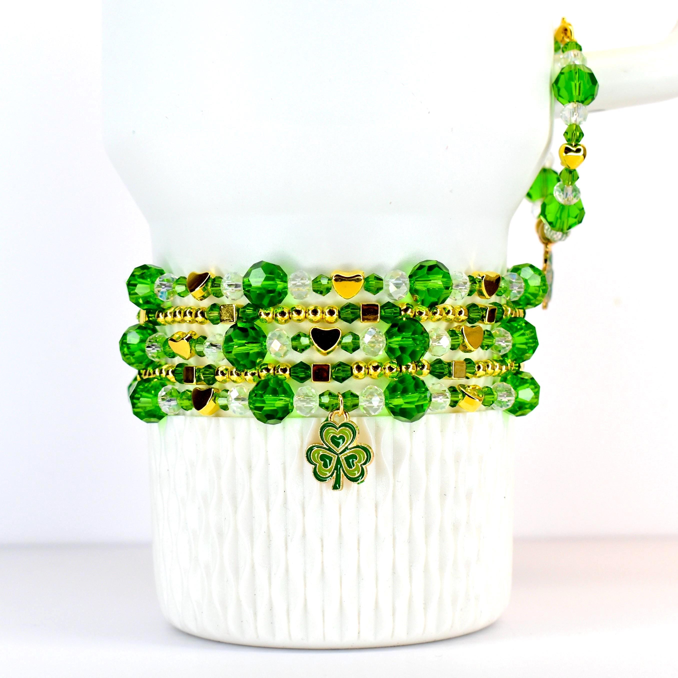 A stack of green beaded bracelets with gold accents and heart charms. A single bracelet with a gold chain and green beads is on the side. A gold shamrock charm hangs from the bottom bracelet. The bracelets are displayed on a white surface.