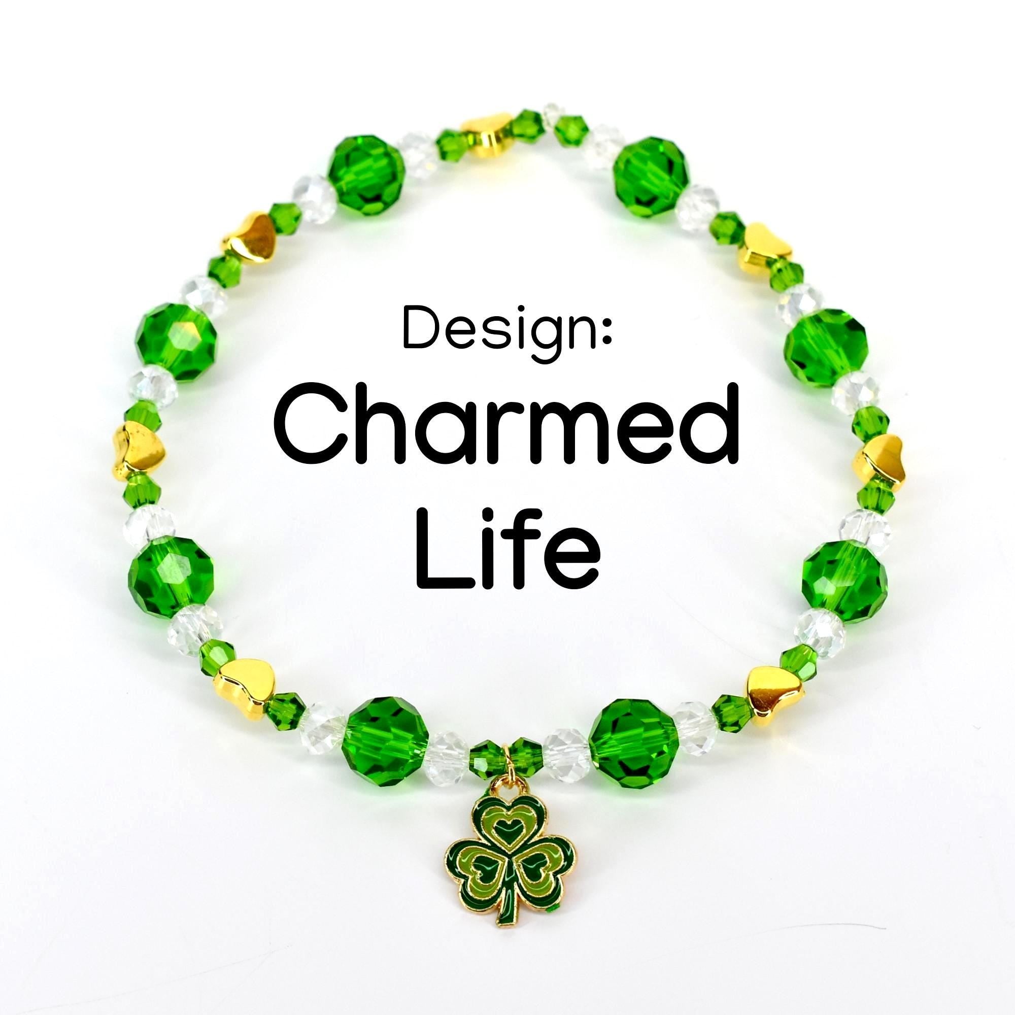 A beaded bracelet with green, clear, and gold beads. The bracelet has a shamrock charm and the text "Design: Charmed Life". The beads are faceted and round, with gold heart-shaped accents.