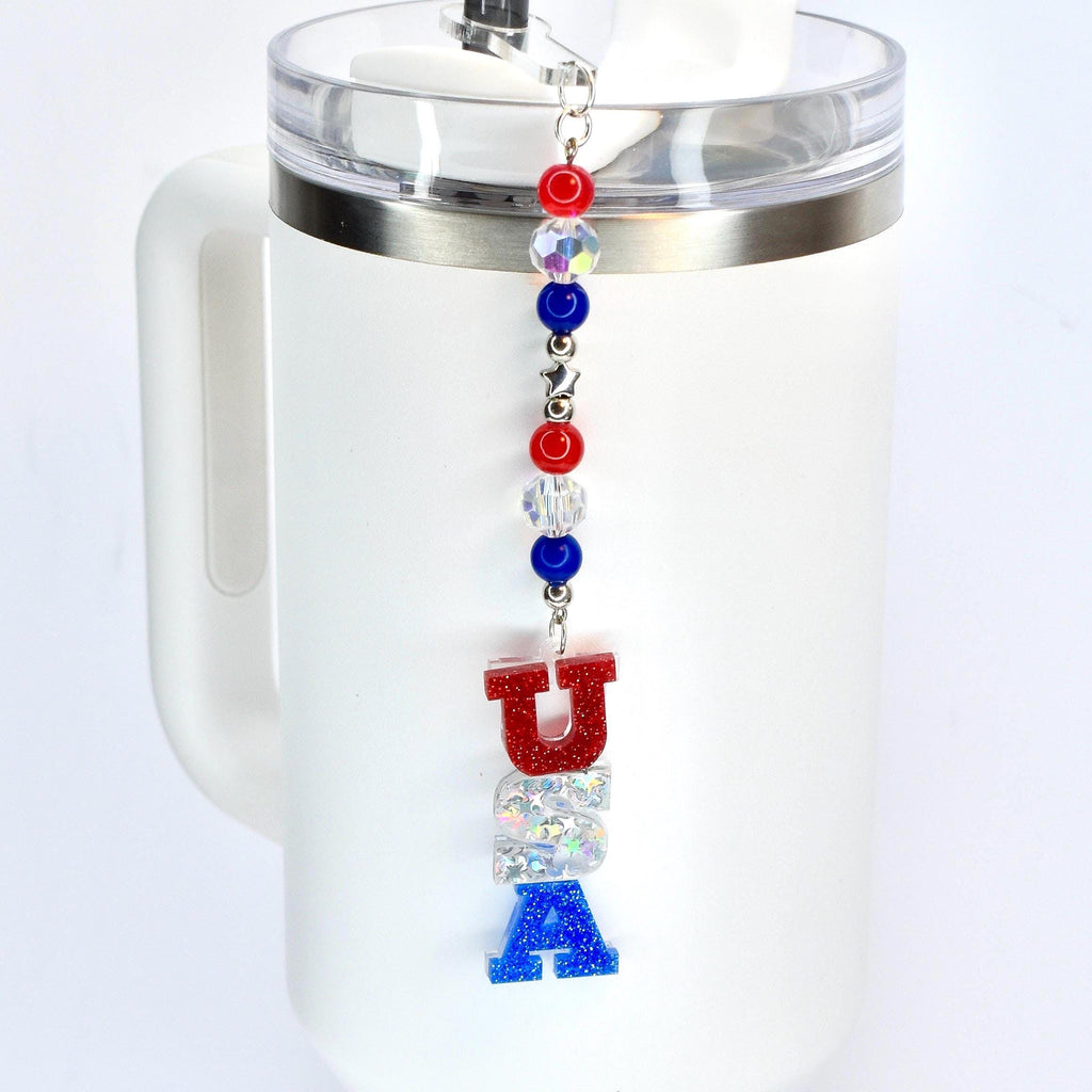 A red white and blue tumbler straw charm with a USA focal charm hanging vertically from the bottom of the charm and red, crystal and blue beads hanging above it.