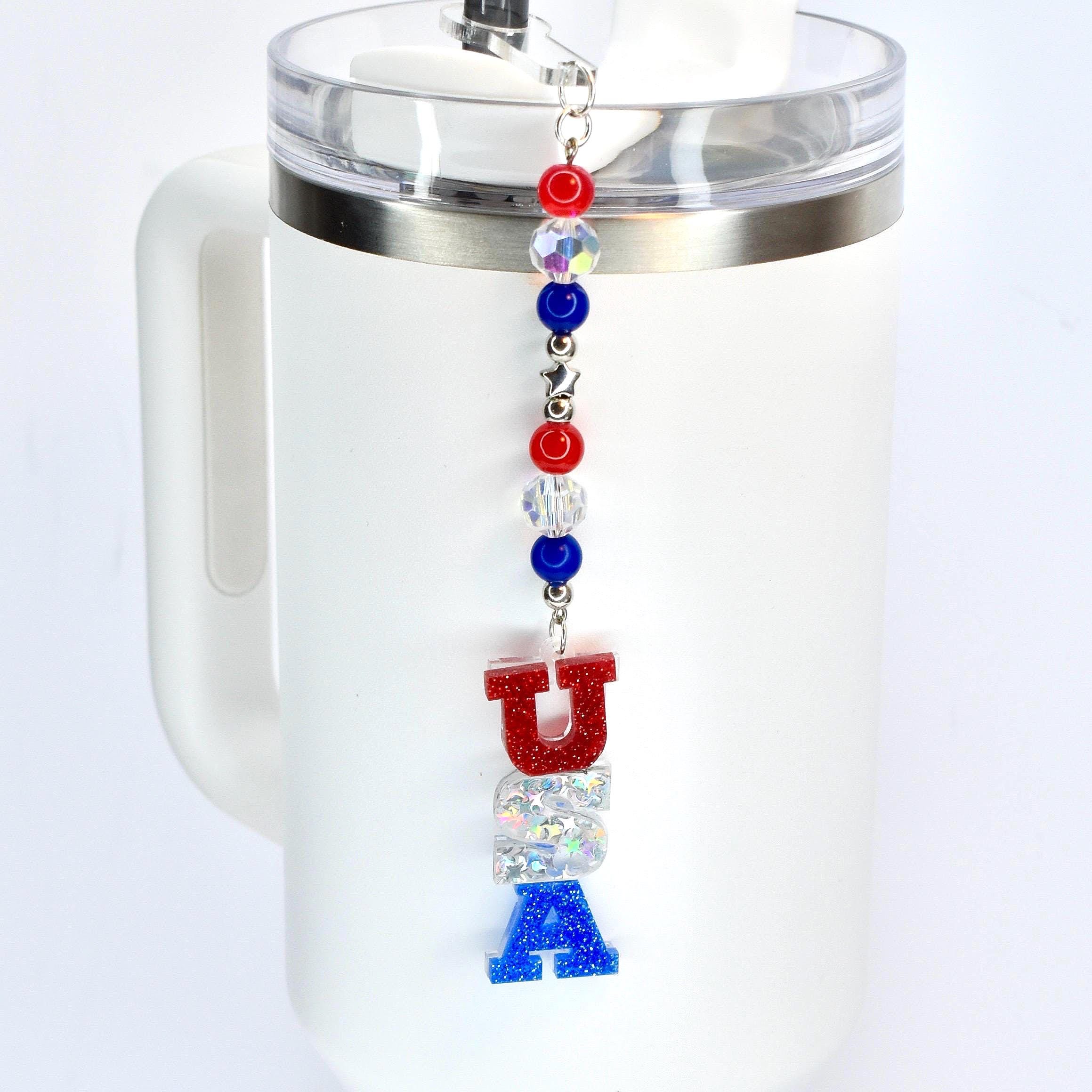 A red white and blue tumbler straw charm with a USA focal charm hanging vertically from the bottom of the charm and red, crystal and blue beads hanging above it.