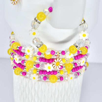 A stack of beaded bracelets in yellow, white, pink, and clear. The bracelets feature daisy and round bead accents. A gold daisy charm hangs from one bracelet. The bracelets are on a white surface.