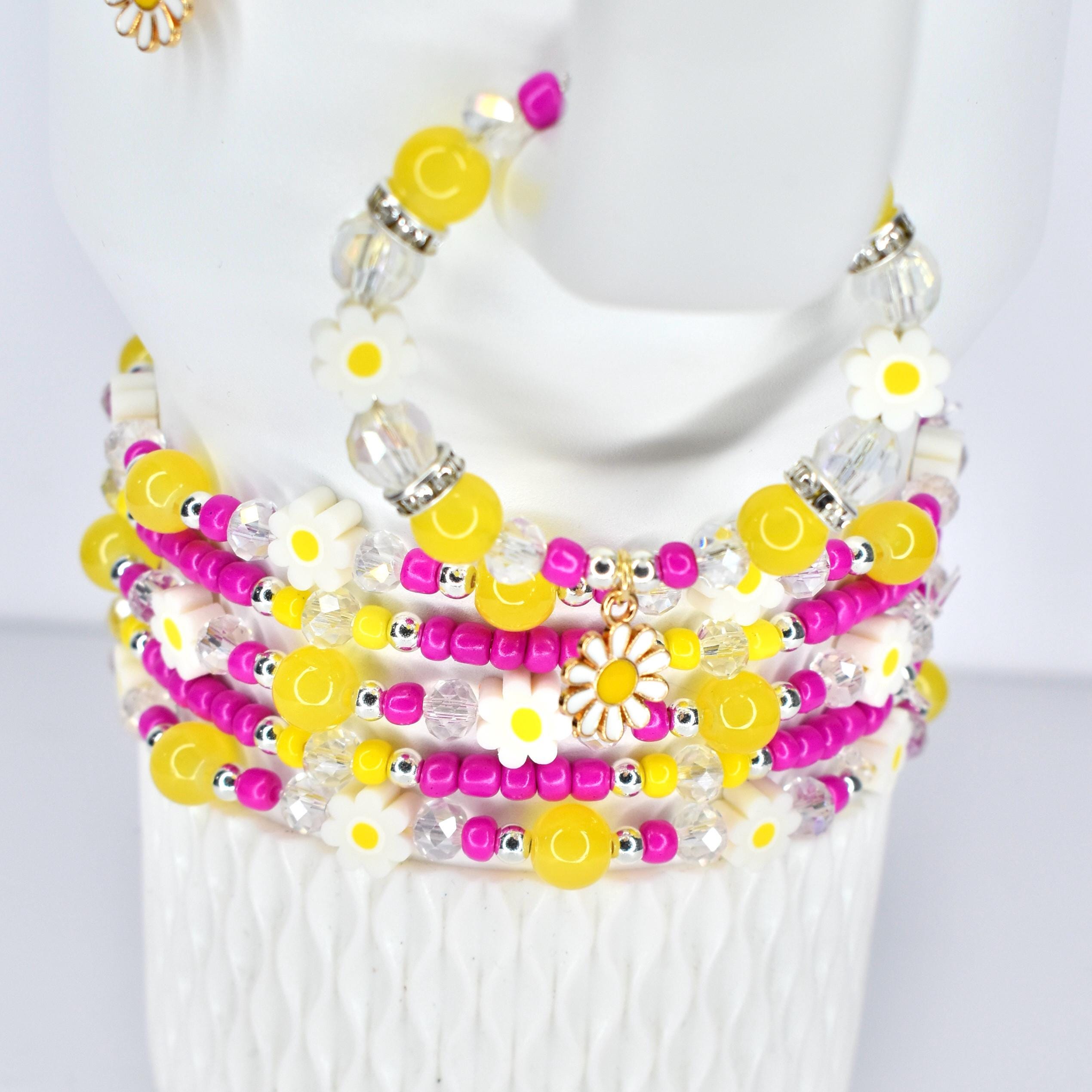 A stack of beaded bracelets in yellow, white, pink, and clear. The bracelets feature daisy and round bead accents. A gold daisy charm hangs from one bracelet. The bracelets are on a white surface.