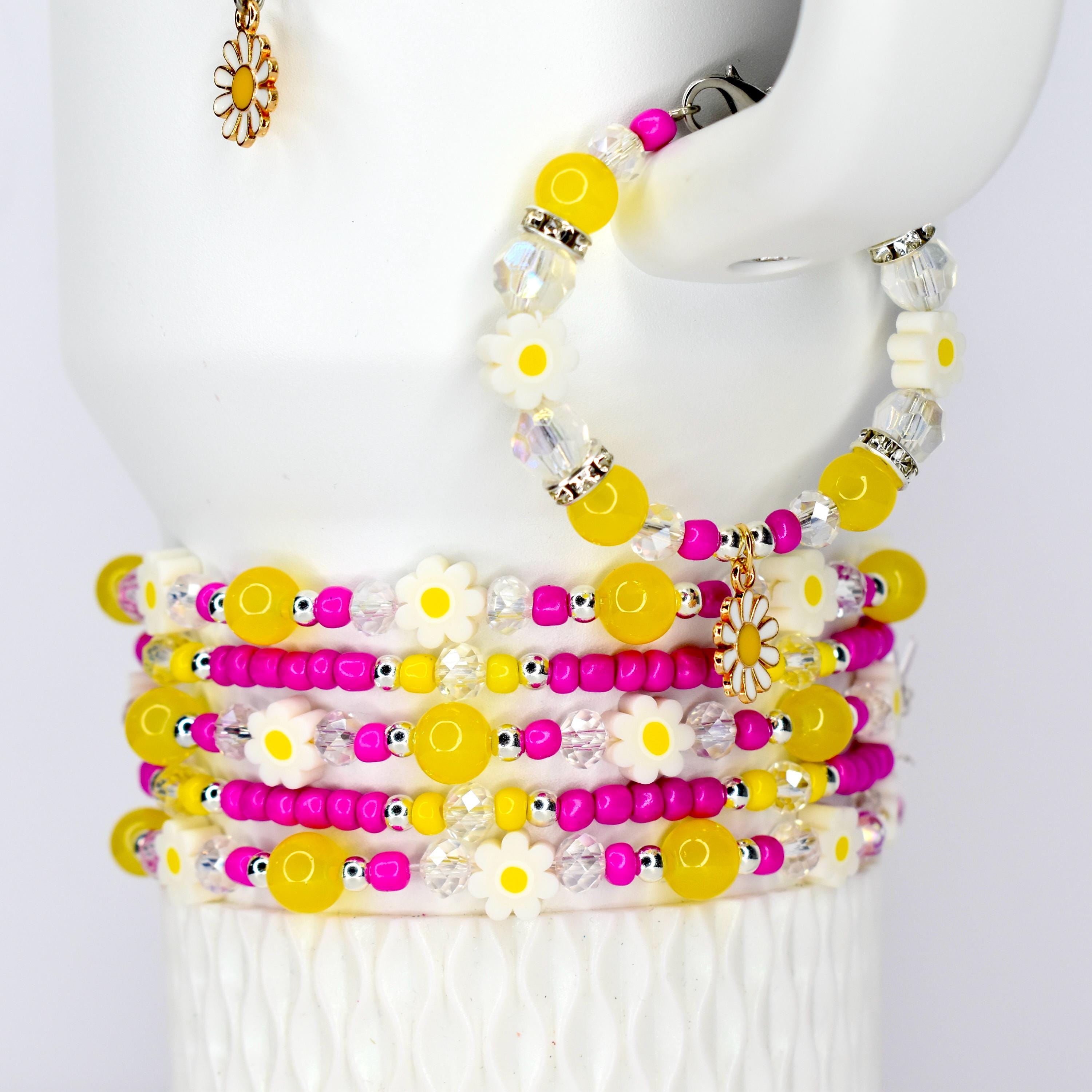 A stack of daisy-themed bracelets with yellow, white, and pink beads. The bracelets are adorned with daisy charms and clear crystal accents. A matching daisy handle charm is also visible.