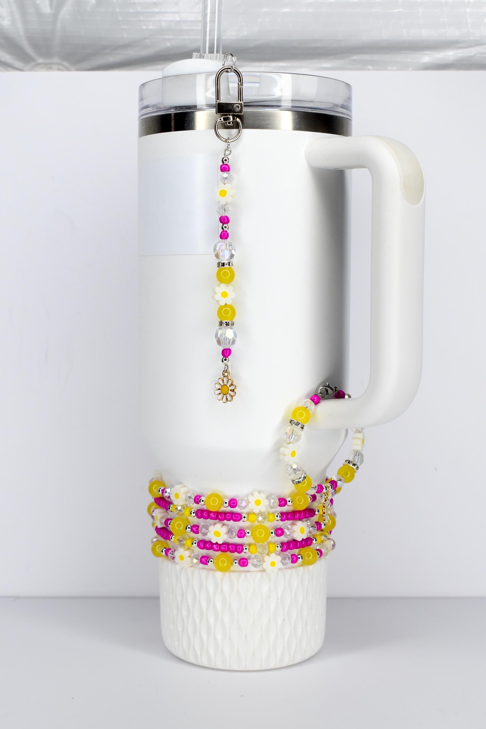 White insulated tumbler with a handle, adorned with a beaded charm and wrapped with beaded bracelets. The charm and bracelets feature yellow, pink, and white daisy and bead designs. A silver clasp secures the charm.