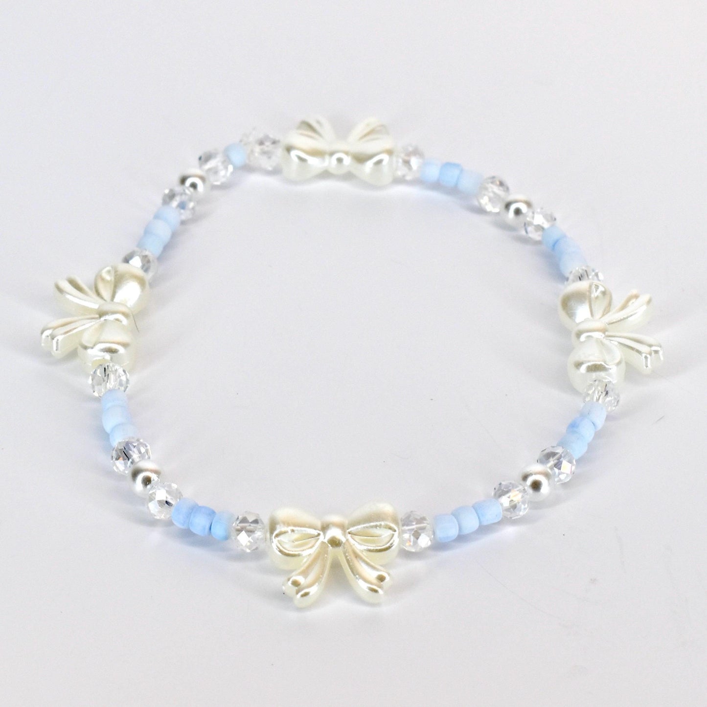 A delicate bracelet featuring a repeating pattern of light blue beads, clear crystal beads, and ivory-colored bow charms. The bracelet is strung on an elastic band, creating a flexible and comfortable fit. The bow charms add a touch of elegance.