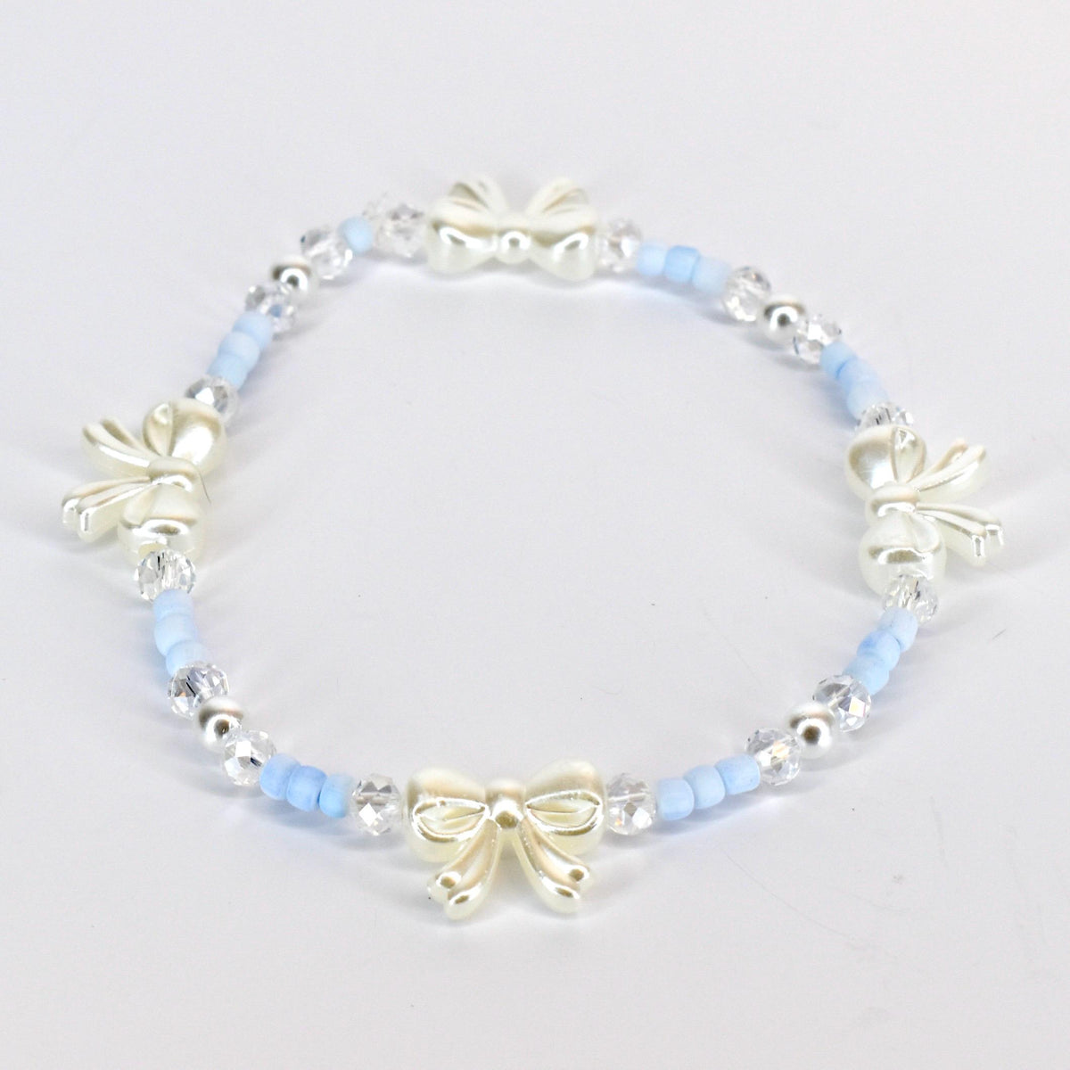 A delicate bracelet featuring a repeating pattern of light blue beads, clear crystal beads, and ivory-colored bow charms. The bracelet is strung on an elastic band, creating a flexible and comfortable fit. The bow charms add a touch of elegance.
