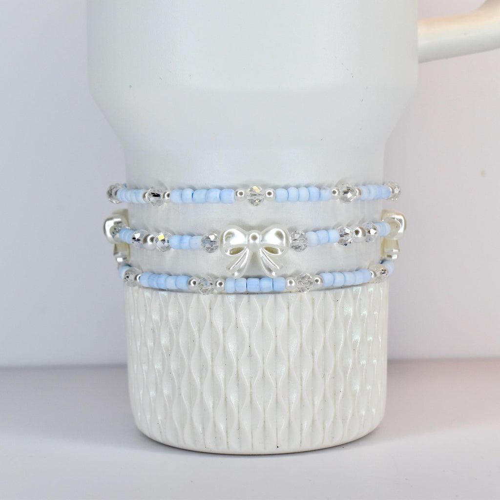 Three light blue beaded bracelets with silver accents and a white bow are stacked on a white ceramic mug. The bracelets have small beads and clear crystal accents. The mug has a textured, ridged design.