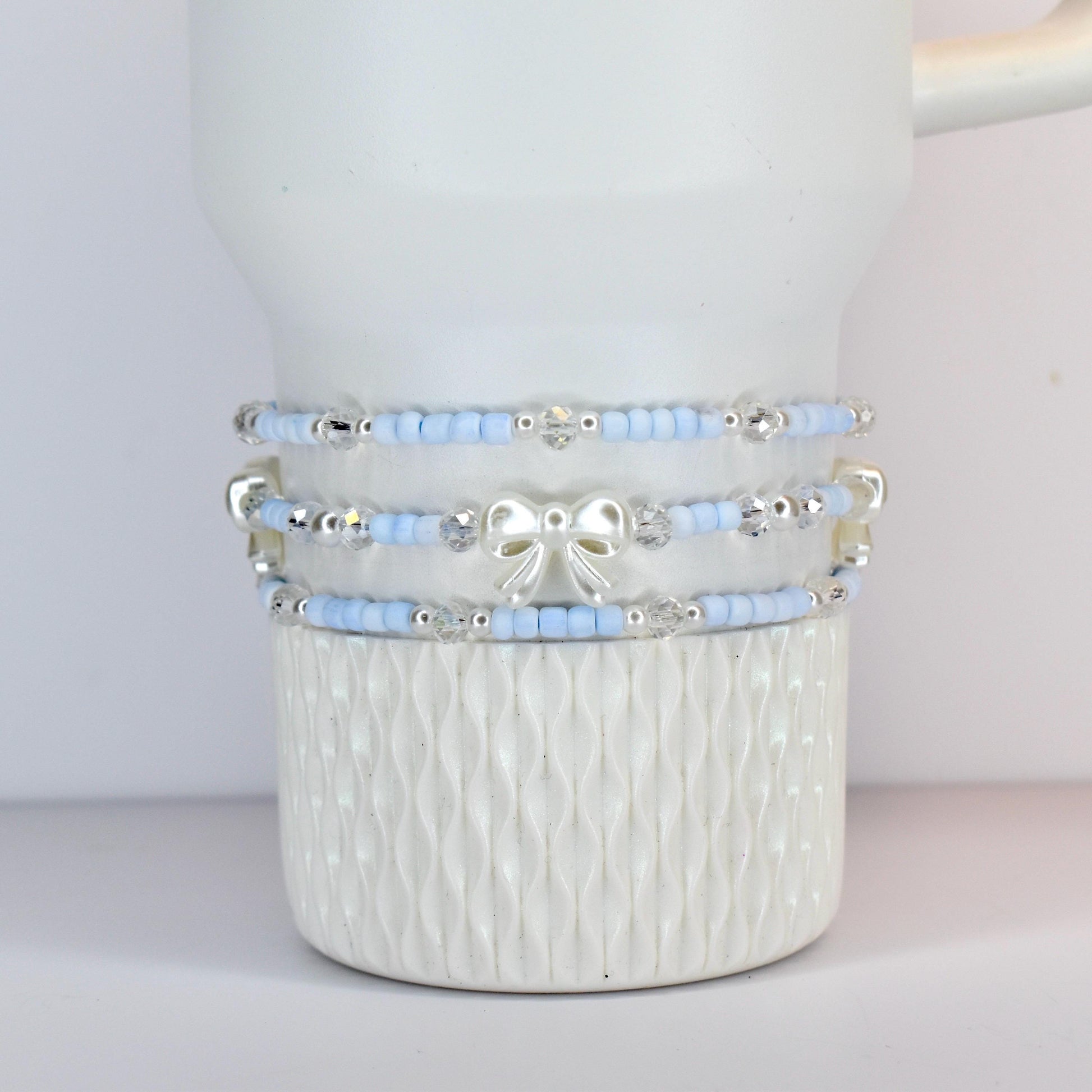 Three light blue beaded bracelets with silver accents and a white bow are stacked on a white ceramic mug. The bracelets have small beads and clear crystal accents. The mug has a textured, ridged design.