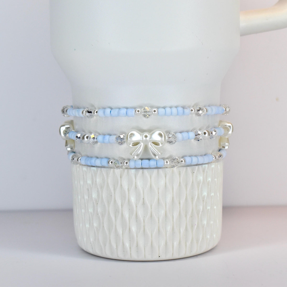 Three light blue beaded bracelets with silver accents and a white bow are stacked on a white ceramic mug. The bracelets have small beads and clear crystal accents. The mug has a textured, ridged design.
