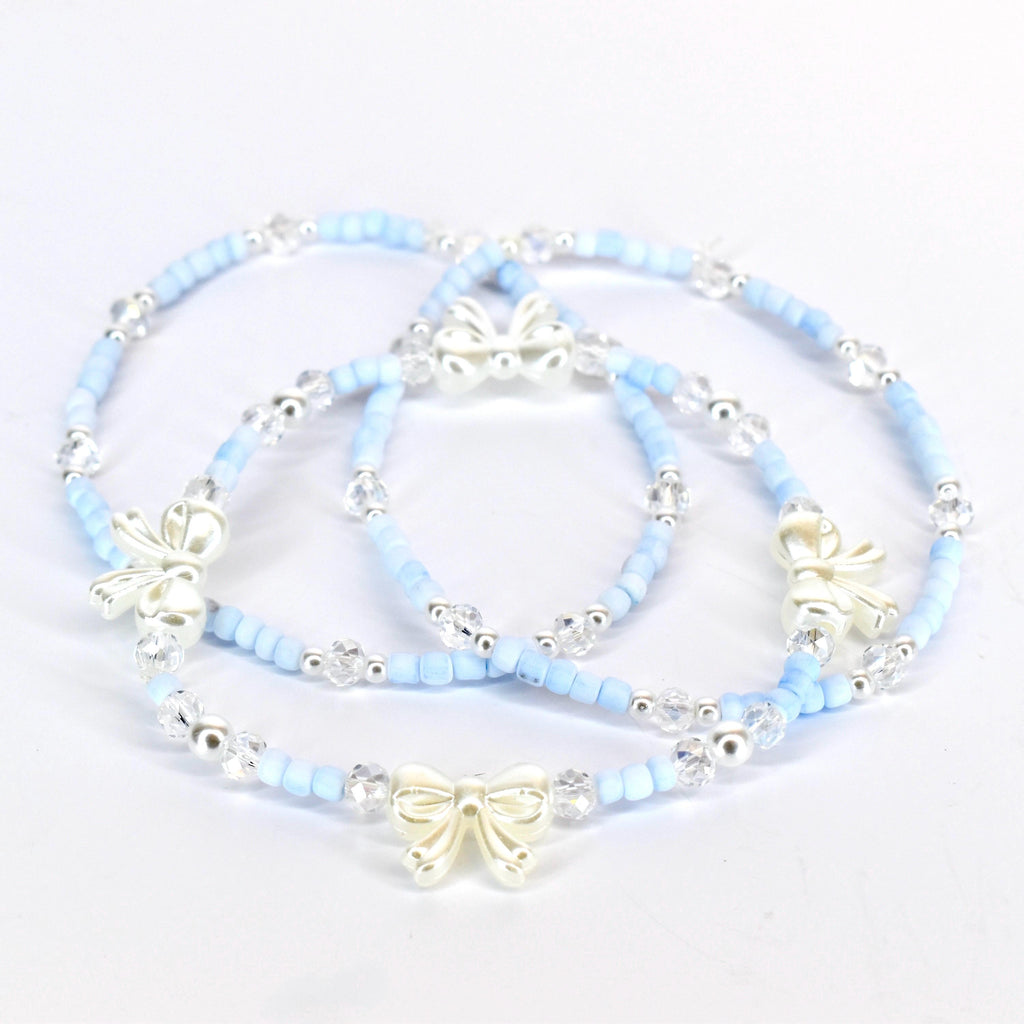 Three delicate bracelets. Each bracelet features light blue beads, clear crystal accents, and a silver-colored bow charm. The bracelets are arranged on a white surface, showcasing their design and details.