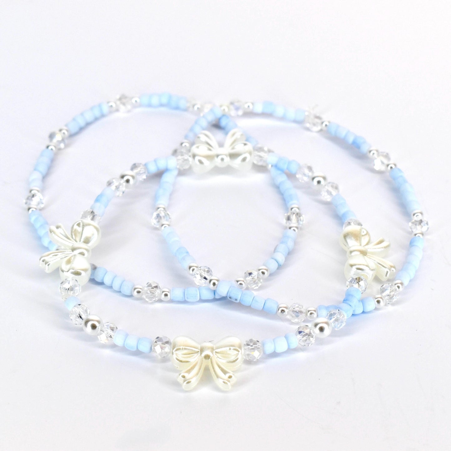 Three delicate bracelets. Each bracelet features light blue beads, clear crystal accents, and a silver-colored bow charm. The bracelets are arranged on a white surface, showcasing their design and details.