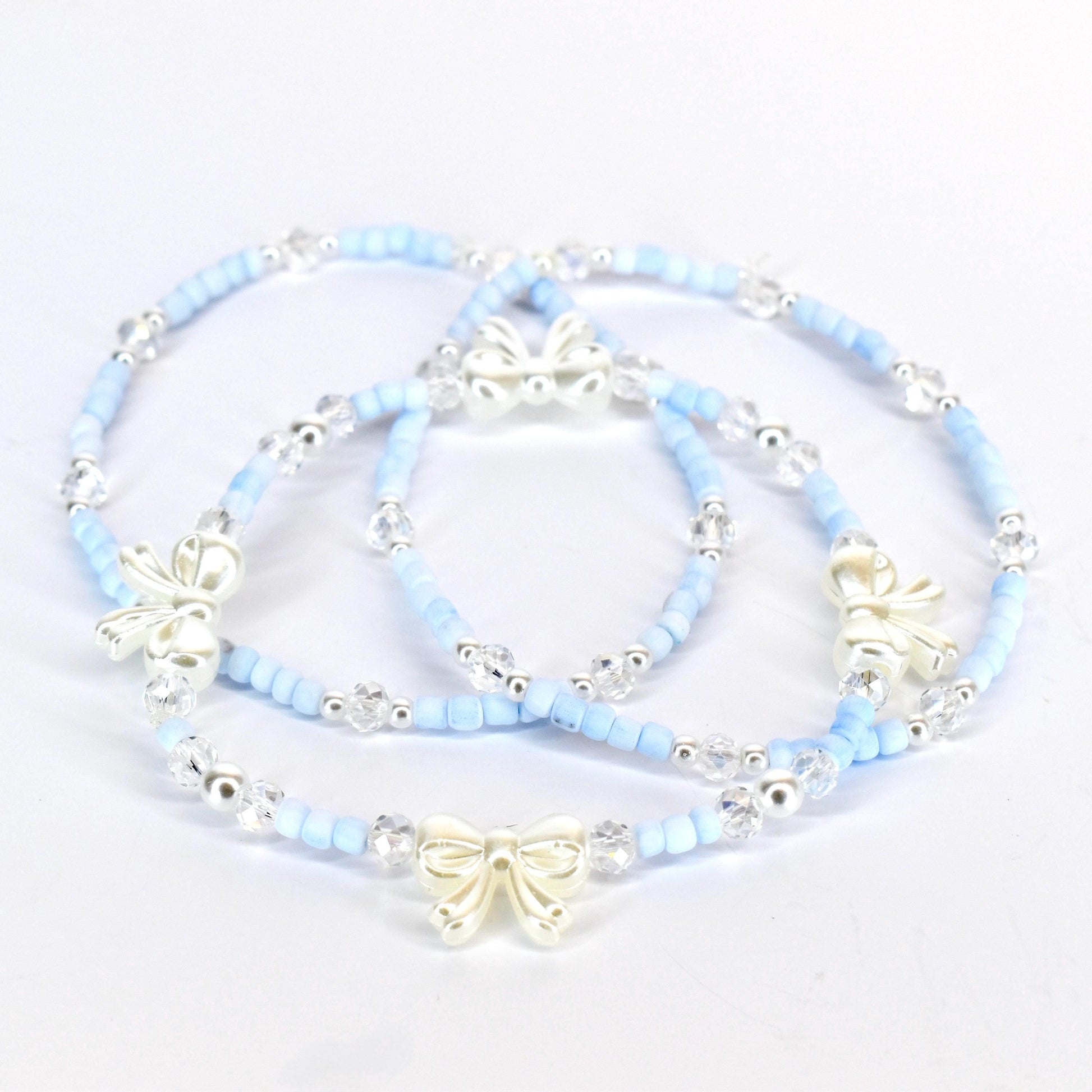 Three delicate bracelets. Each bracelet features light blue beads, clear crystal accents, and a silver-colored bow charm. The bracelets are arranged on a white surface, showcasing their design and details.