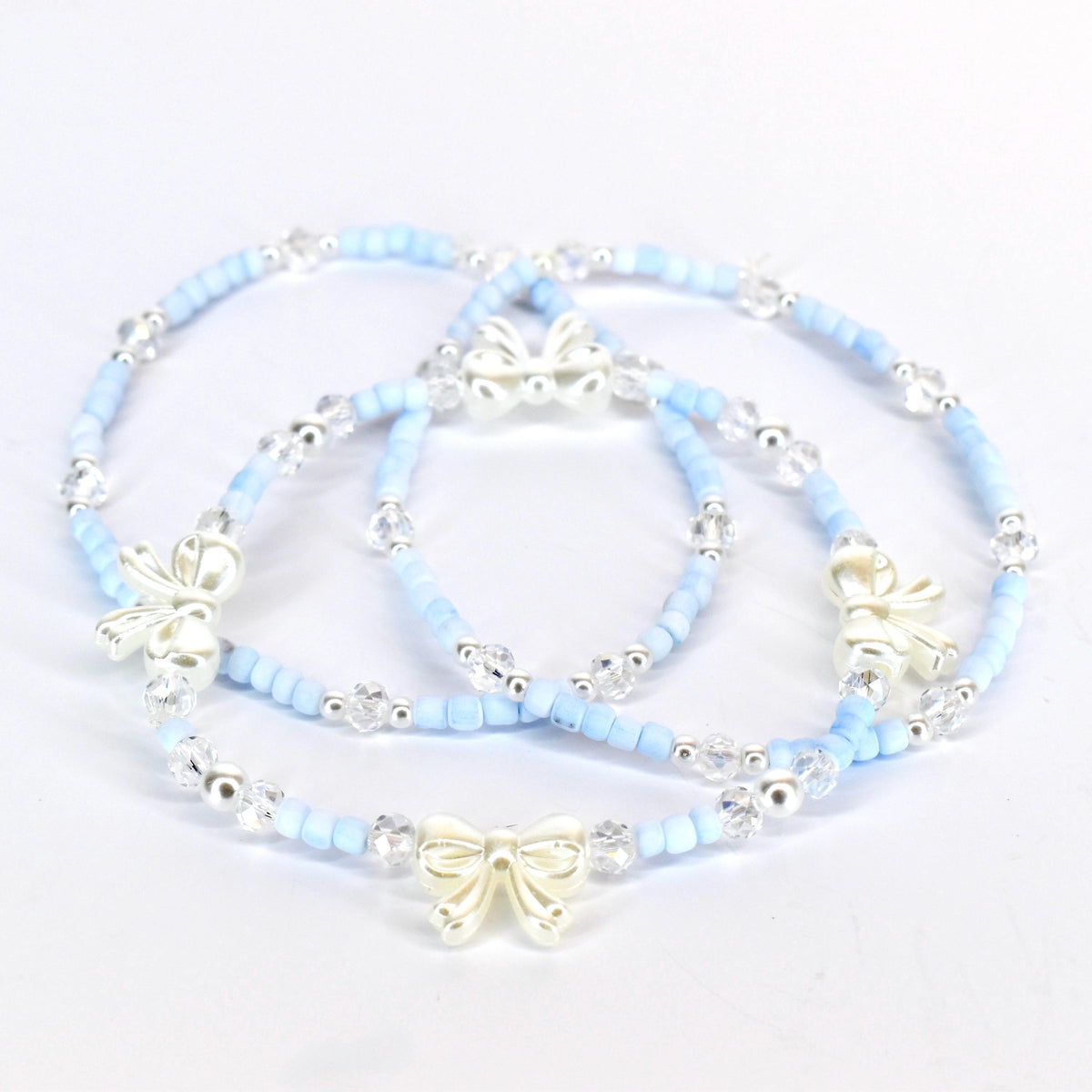Three delicate bracelets. Each bracelet features light blue beads, clear crystal accents, and a silver-colored bow charm. The bracelets are arranged on a white surface, showcasing their design and details.