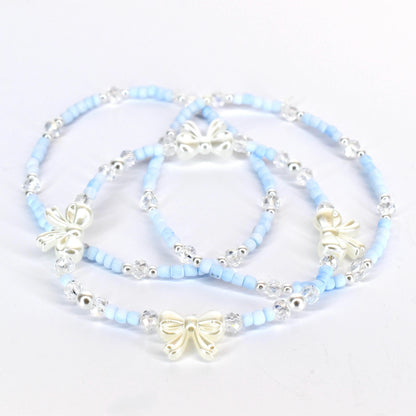 Three delicate bracelets. Each bracelet features light blue beads, clear crystal accents, and a silver-colored bow charm. The bracelets are arranged on a white surface, showcasing their design and details.