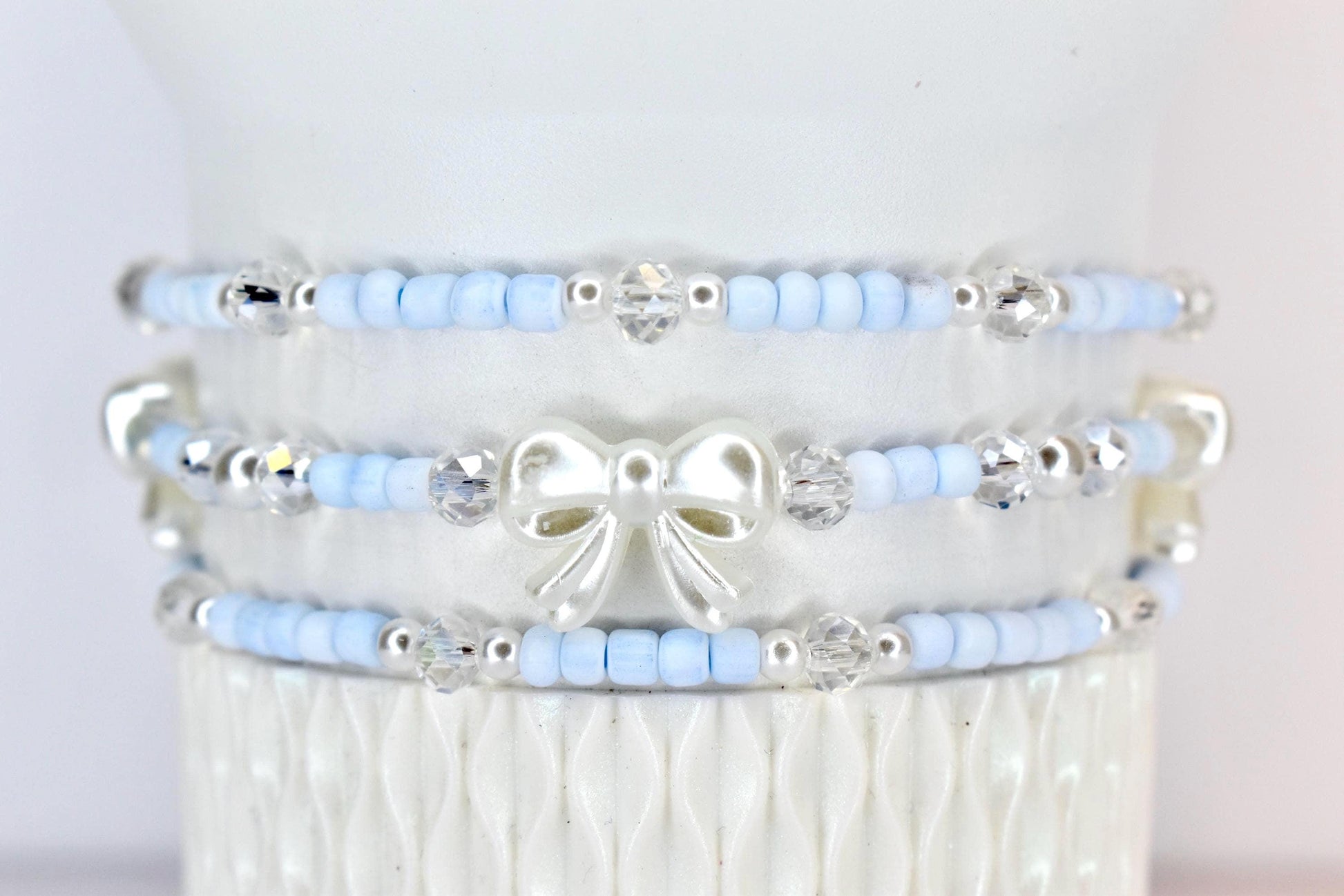 Three beaded bracelets. Each bracelet features light blue beads, silver accents, and clear crystal beads. The middle bracelet has a white bow charm. The bracelets are stacked on a white surface.