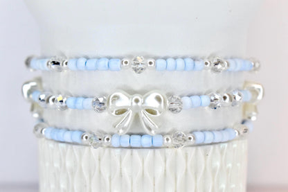 Three beaded bracelets. Each bracelet features light blue beads, silver accents, and clear crystal beads. The middle bracelet has a white bow charm. The bracelets are stacked on a white surface.