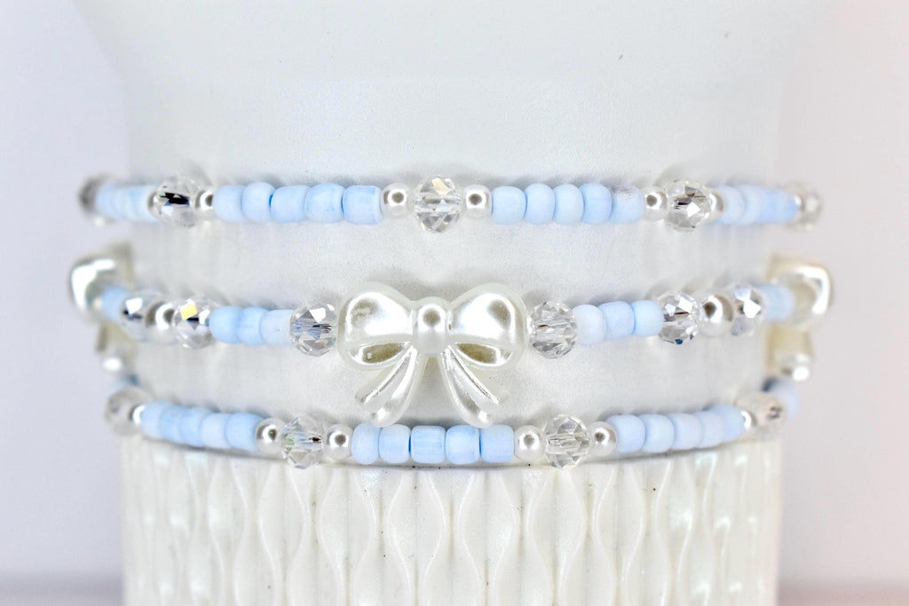 Three beaded bracelets. Each bracelet features light blue beads, silver accents, and clear crystal beads. The middle bracelet has a white bow charm. The bracelets are stacked on a white surface.