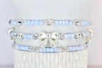 Three beaded bracelets. Each bracelet features light blue beads, silver accents, and clear crystal beads. The middle bracelet has a white bow charm. The bracelets are stacked on a white surface.